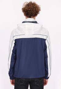 Marineblauwe en witte windjack met capuchon, voorzien van contrasterende zijpanelen en elastische manchetten, geweven stof en een gladde textuur.