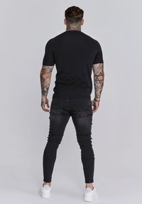 Fekete rövid ujjú ing, sötét skinny farmerral és fehér sportcipővel párosítva. Az öltözék testhez simuló kialakítással és minimális díszítéssel rendelkezik.