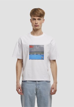Weißes Baumwoll-T-Shirt mit einem quadratischen Motiv und dem Schriftzug "blur" in Rot, das eine blaue Ozeanszene mit Wolken und einem einzelnen Stuhl zeigt.