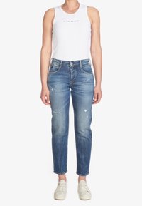 White ribbed tank top with logo positioned above blue distressed jeans. Jeans feature a straight cut and subtle fading, paired with light sneakers.