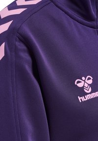 Gros plan d'un maillot de sport violet avec le logo hummel rose et un motif chevron sur l'épaule.