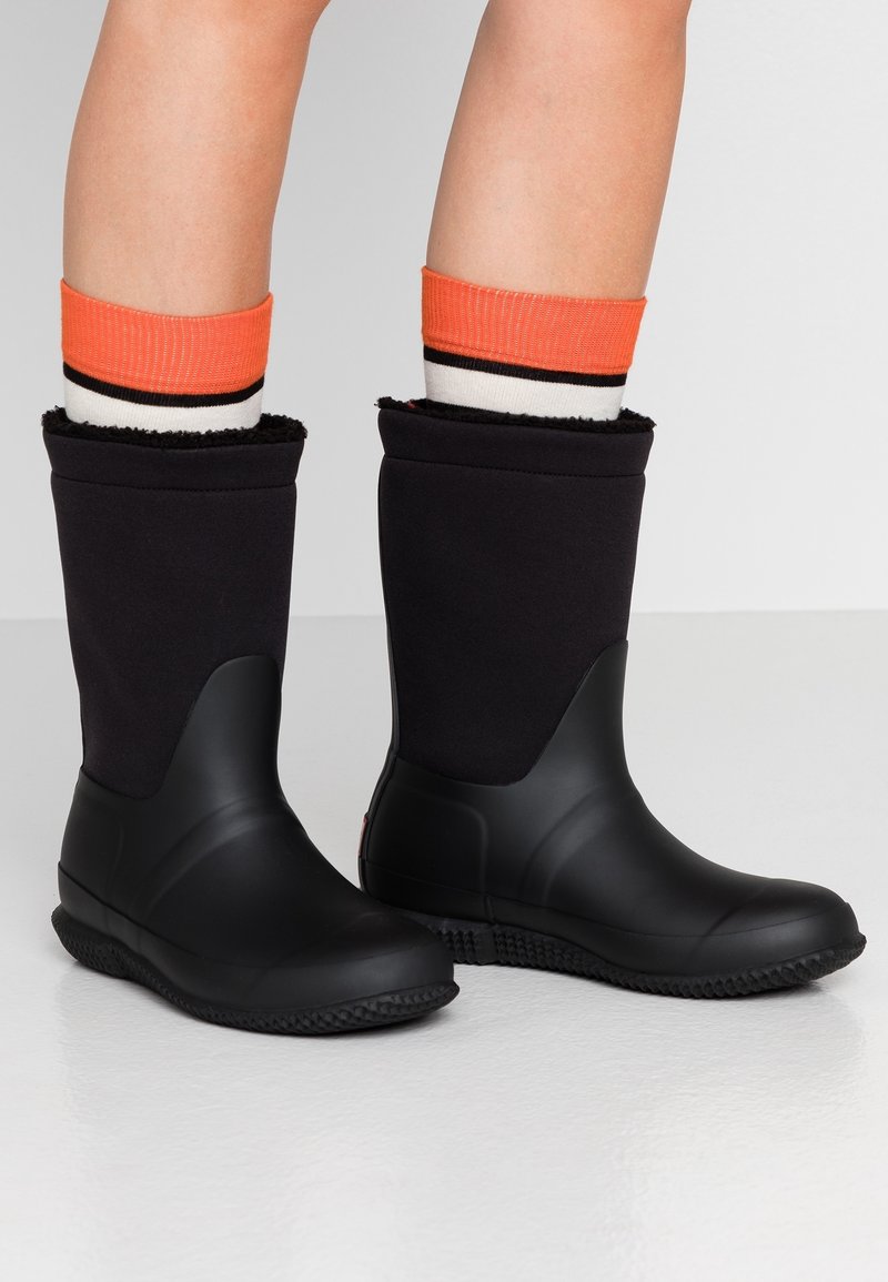 Bottes en caoutchouc noires mi-mollet portées avec des chaussettes rayées orange et blanc sur des jambes nues, debout sur un sol blanc brillant.