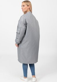Vrouw die met haar rug naar de camera staat in een lange grijze bomberjack, blauwe spijkerbroeken en witte sneakers op een witte achtergrond.
