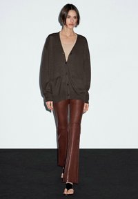 Tmavě hnědý kardigan s výstřihem do V a knoflíkovým zapínáním, s oversized rukávy a dvěma předními kapsami, ve spojení s hnědými rozšířenými kalhotami.