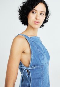 Denim jurk in lichtblauw met een mouwloos ontwerp, zijstrikjes, een ronde halslijn en een textuurafwerking. Getailleerde pasvorm met zichtbare stiksels.