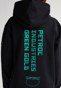 Sudadera negra con texto en negrita color verde azulado: "PETROL INDUSTRIES GREEN GOLD." Incluye logo y detalles de ubicación en la parte inferior. Tejido suave, diseño con capucha.