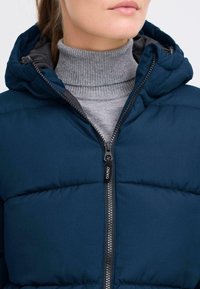 Mornarsko modra napihnjena jakna z zadrgo in kapuco. Ima kvačkan vzorec in sivo rebrasto turtleneck majico spodaj.