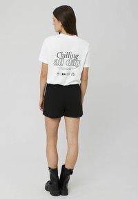 T-shirt blanc à manches courtes avec un imprimé graphique au dos qui indique "Se détendre toute la journée", associé à un short noir et des bottes noires imposantes.