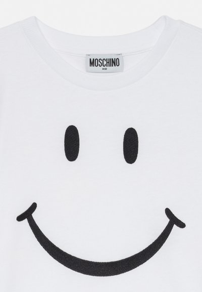 Bela otroška majica s velikim črnim izvezenim nasmeškom in oznako znamke Moschino na ovratniku.