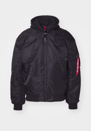 Veste bomber noire avec capuche amovible, fermeture éclair, poignets côtelés et poches latérales. Présente une étiquette rouge sur la manche pour le détail.