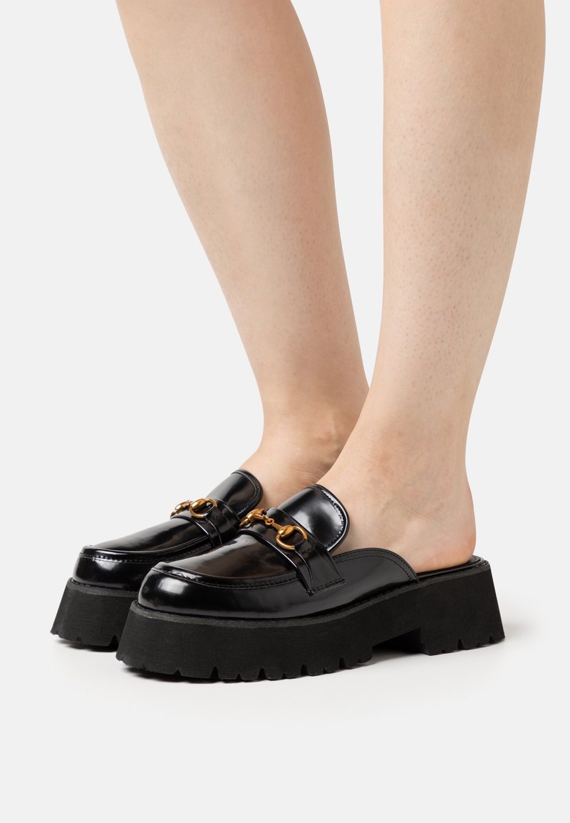 Mocassins slip-on en cuir verni noir avec semelles compensées épaisses et détail en métal doré en forme de mors, portés pieds nus.