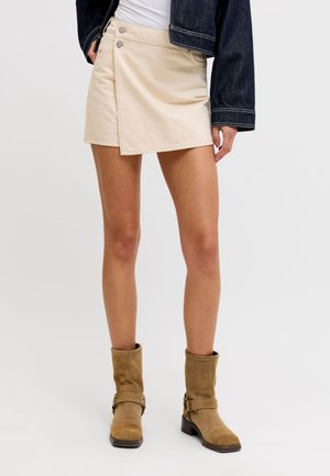 Kvinne iført beige asymmetrisk miniskjørt, brune semsket skinn ankelstøvler, hvit topp og mørk denimjakke, står mot en ensfarget bakgrunn.