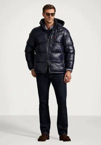 Mies, joka käyttää mustaa untuvatakkia, tummia farkkuja, ruskeita kenkiä, aurinkolaseja ja kelloa, seisoo tasaista vaaleaa taustaa vasten.