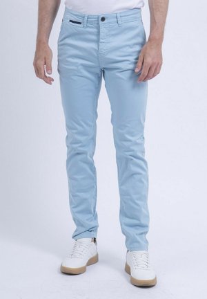 Homme portant un pantalon slim bleu clair et des baskets blanches, debout devant un fond uni.