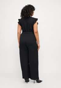 Black blouse with eyelet detailing and ruffled sleeves, paired with wide-legged black pants and heeled black shoes. Back view.