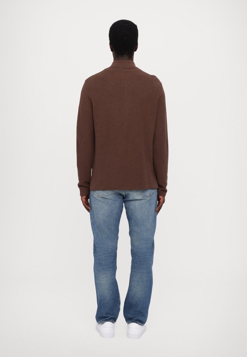 Braunes Strickoberteil mit langen Ärmeln und Kragen, kombiniert mit hellblauen Jeans. Weiße Sneakers runden das Outfit ab. Ansicht von hinten.