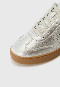 Baskets argentées métalliques avec lacets beige et semelle en caoutchouc marron, en cuir texturé avec détail décoratif en strass sur le côté.