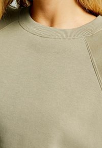 Ljus olivgrön sweatshirt med ribbad rundhals, texturerad tyg och raglanärmar. Enkelt design, inga synliga logotyper eller mönster.