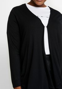 Osoba ubrana w czarny długi kardigan z długim rękawem na białą koszulę z częściowym nadrukowanym tekstem, ukazana od ramion do talii.