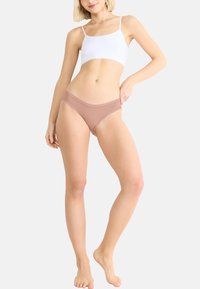 Beżowa bielizna hipsterska z wyszukanym koronkowym wykończeniem i logo na gumce, zestawiona z gładkim białym topem na ramiączkach.