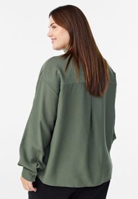 Kobieta z długimi brązowymi włosami, ubrana w luźną, oliwkowozieloną bluzkę z długim rękawem, z boku i uśmiechająca się na białym tle.