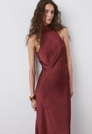 Ženska z dolgimi kodrastimi lasmi, oblečena v brezrokavno bordo halterneck obleko, stoji ob preprosti beli steni z dvema zlatima zapestnicama.