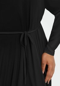 Robe noire à manches longues, dotée d'une ceinture nouée à la taille. Tissu lisse au texture fluide, soulignant la silhouette.