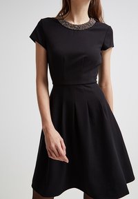 Schwarzes Kleid mit einem figurbetonten Oberteil und einem schwingenden Rock, ausgestattet mit einem mit Perlen verzierten Ausschnitt und kurzen Ärmeln. Die glatte Stofftextur betont die Eleganz.