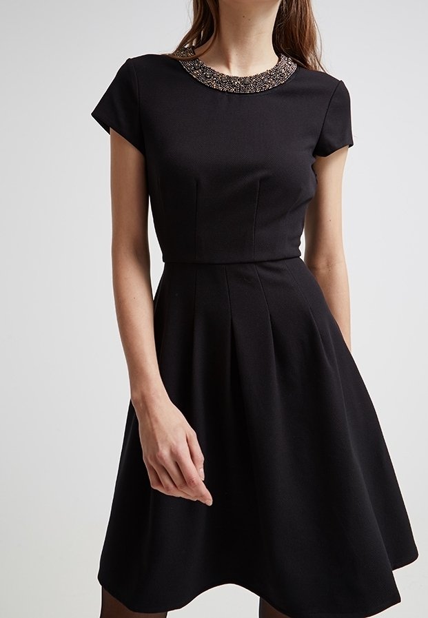 Schwarzes Kleid mit einem figurbetonten Oberteil und einem schwingenden Rock, ausgestattet mit einem mit Perlen verzierten Ausschnitt und kurzen Ärmeln. Die glatte Stofftextur betont die Eleganz.