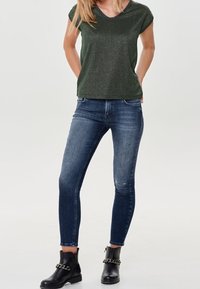 Mörkgrön kortärmad topp med V-hals, parat med mörkblå skinny jeans med en blekt textur och små framknäpatcher, kompletterat med svarta ankelskor med kedjor.