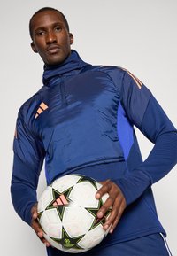 Granatowa i niebieska sportowa kurtka z wysokim kołnierzem, ozdobiona pomarańczowymi paskami i zamkiem. Model trzyma białą i czarną piłkę nożną z zielonymi akcentami.