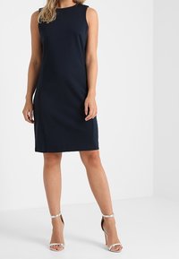 Granatowa sukienka bez rękawów z gładkiego materiału, o prostym kroju i długości do kolan, zestawiona z srebrnymi sandałami na obcasie.