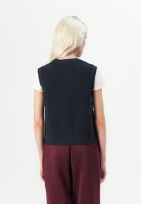 Marineblauwe gebreide mouwloze trui met een ribbelstructuur, ronde halslijn en een aansluitende taille, gedragen over een witte t-shirt met korte mouwen.