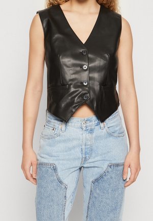Ærmeløs sort lædervest med knapper og V-udskæring, båret over lyseblå denimjeans med store laplommer.