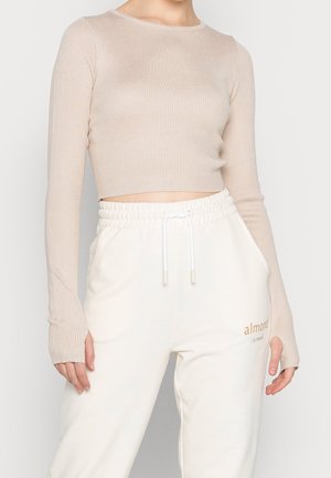 Person iført en beige ribstrikket langærmet crop top og hvide joggers med snøre og teksten "almond" på venstre lår.
