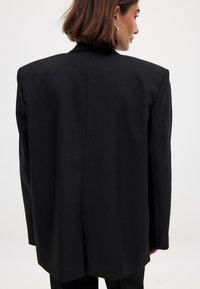 Zwarte oversized blazer met gestructureerde schouders, een gladde textuur en een enkele achterkantopening, die een schoon en minimalistisch design laat zien.