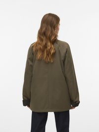 Kobieta z długimi falującymi brązowymi włosami, ubrana w obszerna oliwkową kurtkę i ciemne spodnie, stoi tyłem na białym tle.