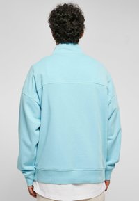 Ljusblå oversized sweatshirt med hög krage, ribbade muddar och en mjuk textur. Har en horisontell söm över ryggen.