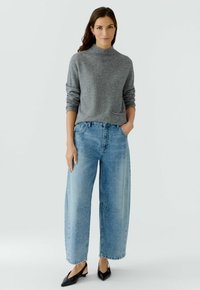 Pull en maille grise avec un col montant, associé à un jean en denim bleu à jambes larges avec des poches avant et un lavage léger. Mocassins noirs à bout pointu.