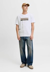 Camiseta blanca de manga corta con un logo gráfico negro y dorado, combinada con jeans ajustados de un tono azul desgastado y calzado beige.
