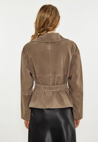 Taupe suède jas met een brede kraag, een ceintuur in de taille en een geplooide achterkant. Zachte textuur en gestructureerde pasvorm, gecombineerd met een zwarte rok.