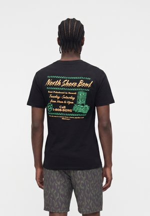 Férfi fonott hajjal, fekete pólót visel, amelyen a North Shore Bowl felirat és hawaii stílusú minta található, mintás rövidnadrággal, háttal a nézőnek.