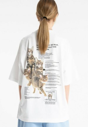 Osoba z długimi włosami związanymi w kucyk, ubrana w białą, oversize'ową koszulkę z wieloma wygładzonymi kotami i nadrukowanym tekstem na plecach.