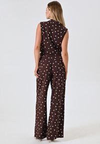 Bruine jumpsuit met polka dots, wijduitlopende pijpen, mouwloos topje en een aansluitende taille. Glad textuur met contrasterende witte stippen overal.