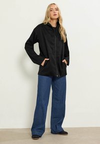 Svart zip-up jacka i mjukt tyg, med hög krage och framfickor. Matchad med vida denimjeans och svarta skor.