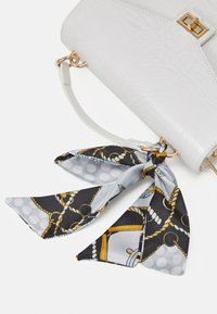 Sac à main blanc texturé avec un détail de fermeture dorée, orné d'un foulard noir et gris à motifs attaché à la poignée. Le foulard présente des motifs de corde et de pois.