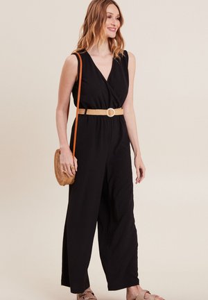 Schwarzer ärmelloser Jumpsuit mit Wickeldesign und weitem Bein, tailliert mit einem beigefarbenen geflochtenen Gürtel. Als Accessoires wird eine runde Strohtasche getragen.
