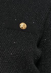 Μαύρο υφαντό ύφασμα με υφή και σχέδιο πλέξης, διακοσμημένο με λαμπερά παγιέτες. Περιλαμβάνει μια μαύρη πλεκτή ταινία με χρυσό κουμπί με ετικέτα "B.C."