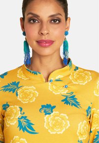 Gelbes Blumenkleid mit blauen und weißen Blumenmustern, einem Stehkragen und blauen Knöpfen. Blau-farbene Perlenquastenohrringe sorgen für einen Farbkontrast.