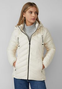 Gevoerde crèmekleurige jas met capuchon, diagonale stiksels en zwarte ritsen; gecombineerd met een lichtgrijze geribbelde trui.